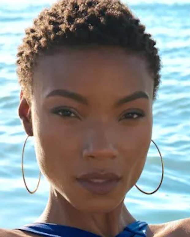 Iyonna Fairbanks poses in front of the ocean in a deep blue bikini and oversized hoop earrings.